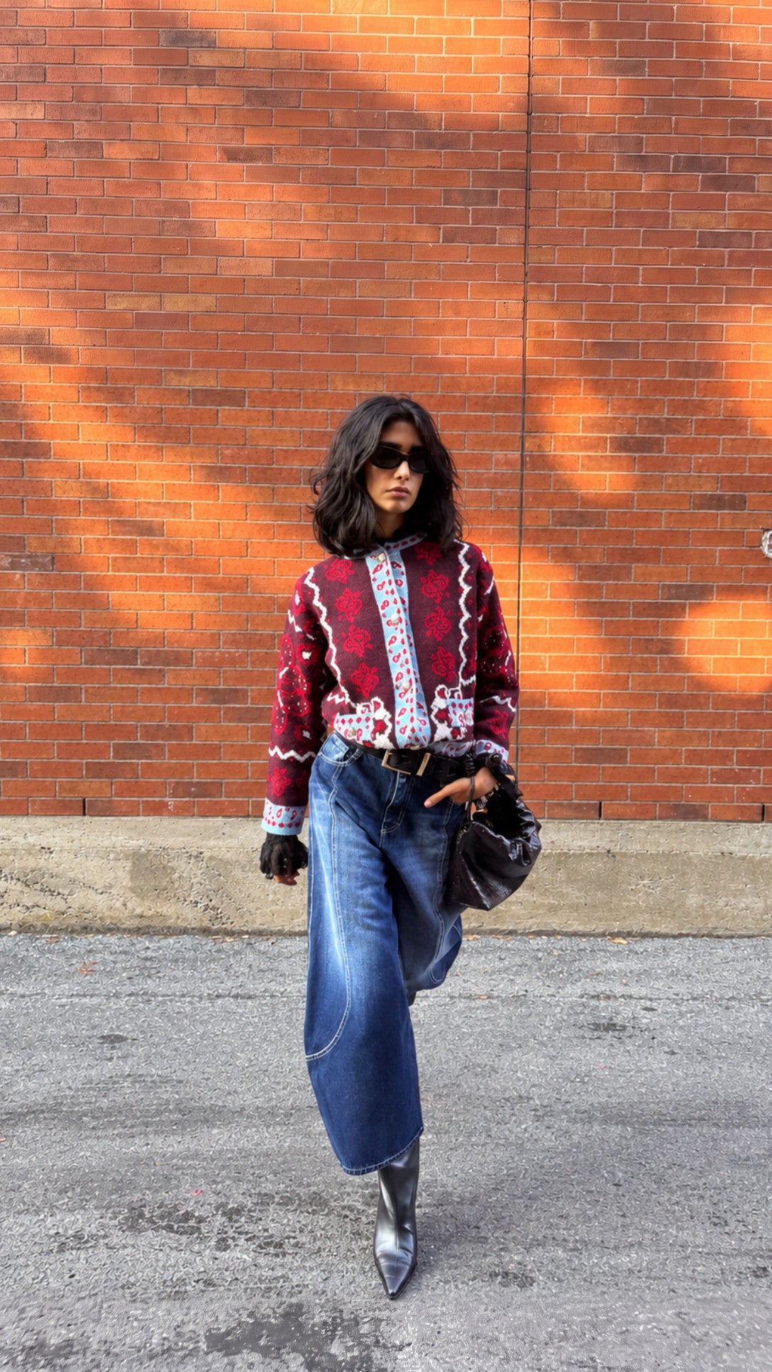 Model in a red print knit cardigan and wide blue jeans walking past a brick wall