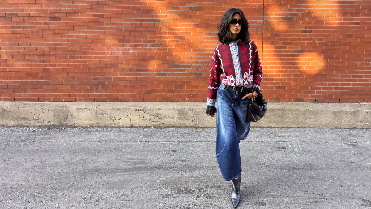 Model in a red print knit cardigan and wide blue jeans walking past a brick wall